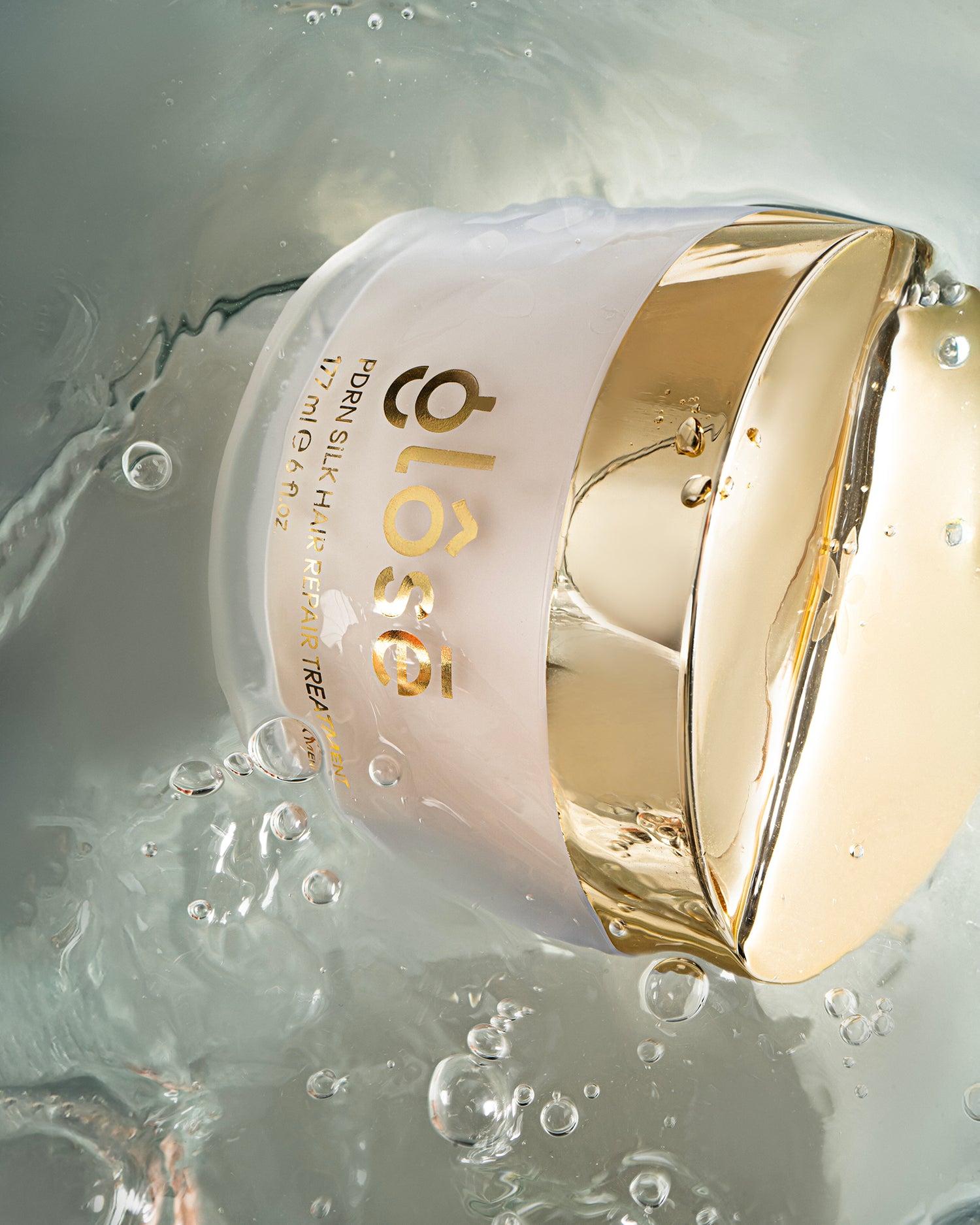 A close-up of Glôsē's PDRN Silk Hair Repair Treatment jar with a reflective gold lid and pale pink body, partially submerged in clear bubbly water. The brand name 'Glôsē is printed in gold on the side.