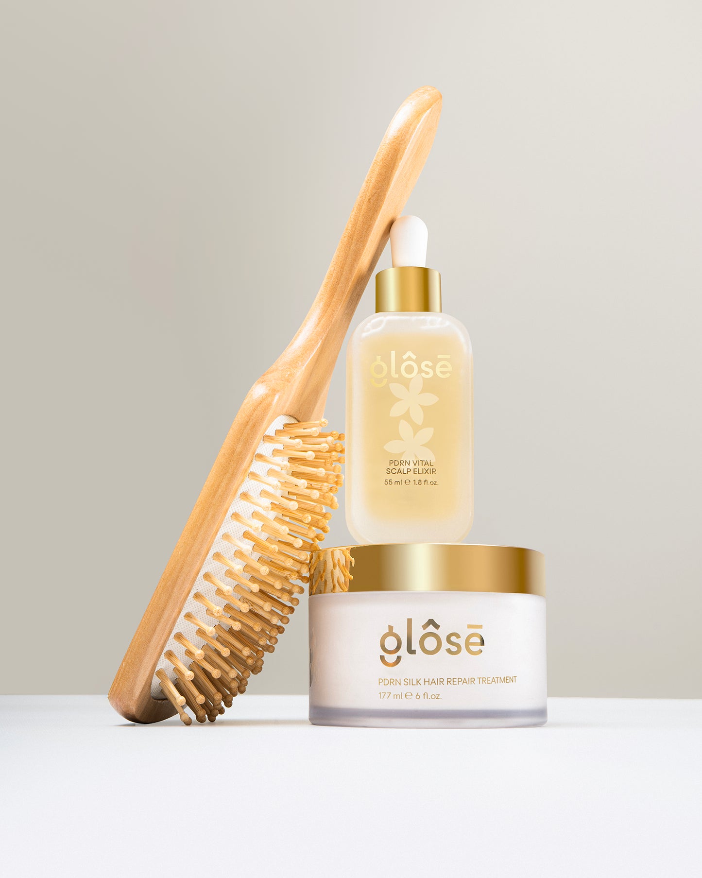 A hair care bundle from Glôsē, featuring a wooden hair brush with natural bristles, a clear bottle of Glôsē's PDRN Vital Scalp Elixir in a glass bottle with a gold cap and white dropper, and a white jar of Glôsē's PDRN Silk Hair Repair Treatment hair mask with a gold lid, arranged on a white surface against a light gray background.
