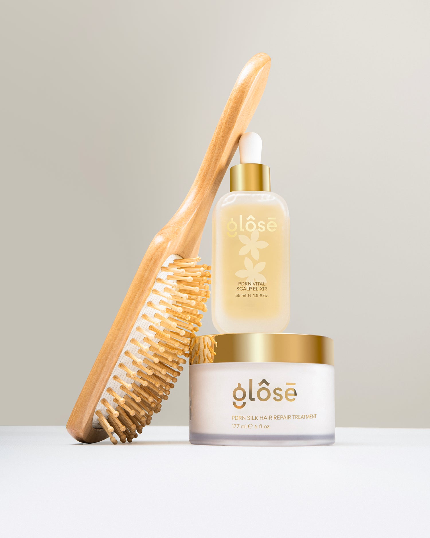 A hair care bundle from Glôsē, featuring a wooden hair brush with natural bristles, a clear bottle of Glôsē's PDRN Vital Scalp Elixir in a glass bottle with a gold cap and white dropper, and a white jar of Glôsē's PDRN Silk Hair Repair Treatment hair mask with a gold lid, arranged on a white surface against a light gray background.
