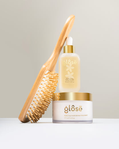 A hair care bundle from Glôsē, featuring a wooden hair brush with natural bristles, a clear bottle of Glôsē's PDRN Vital Scalp Elixir in a glass bottle with a gold cap and white dropper, and a white jar of Glôsē's PDRN Silk Hair Repair Treatment hair mask with a gold lid, arranged on a white surface against a light gray background.