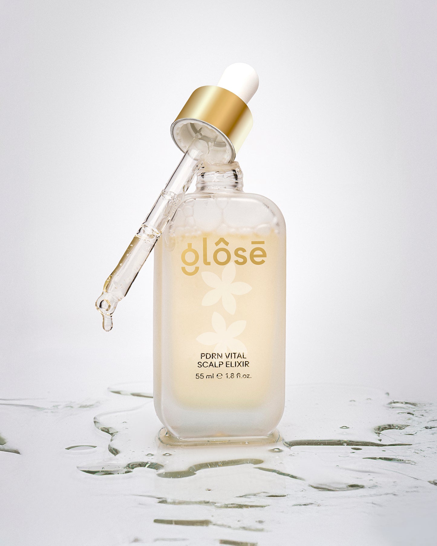 A frosted glass dropper bottle of Glôsē PDRN Vital Scalp Elixir hair serum with a gold collar and white dropper cap, set on a reflective white surface with water droplets.