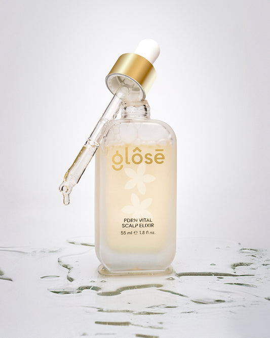 A frosted glass dropper bottle of Glôsē PDRN Vital Scalp Elixir hair serum with a gold collar and white dropper cap, set on a reflective white surface with water droplets.